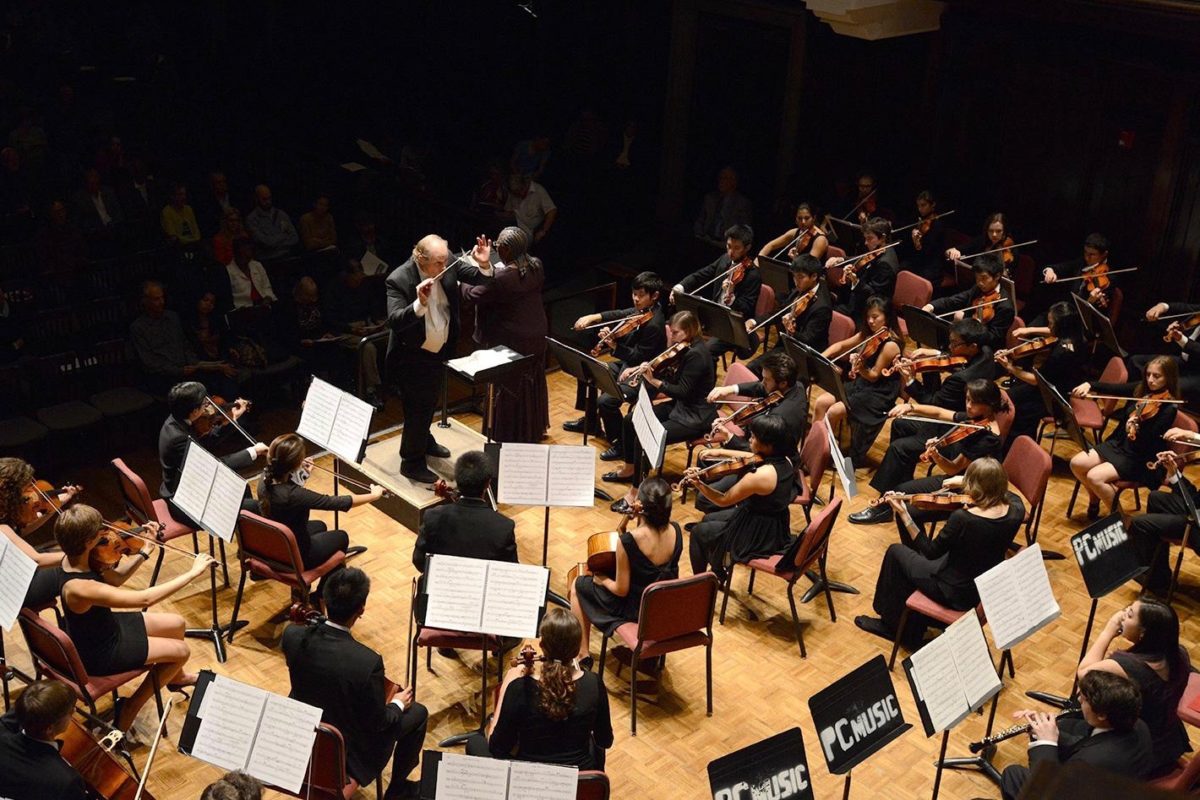 Orchestra performance. Several students sitting in chairs playing instruments directed by a music conductor.