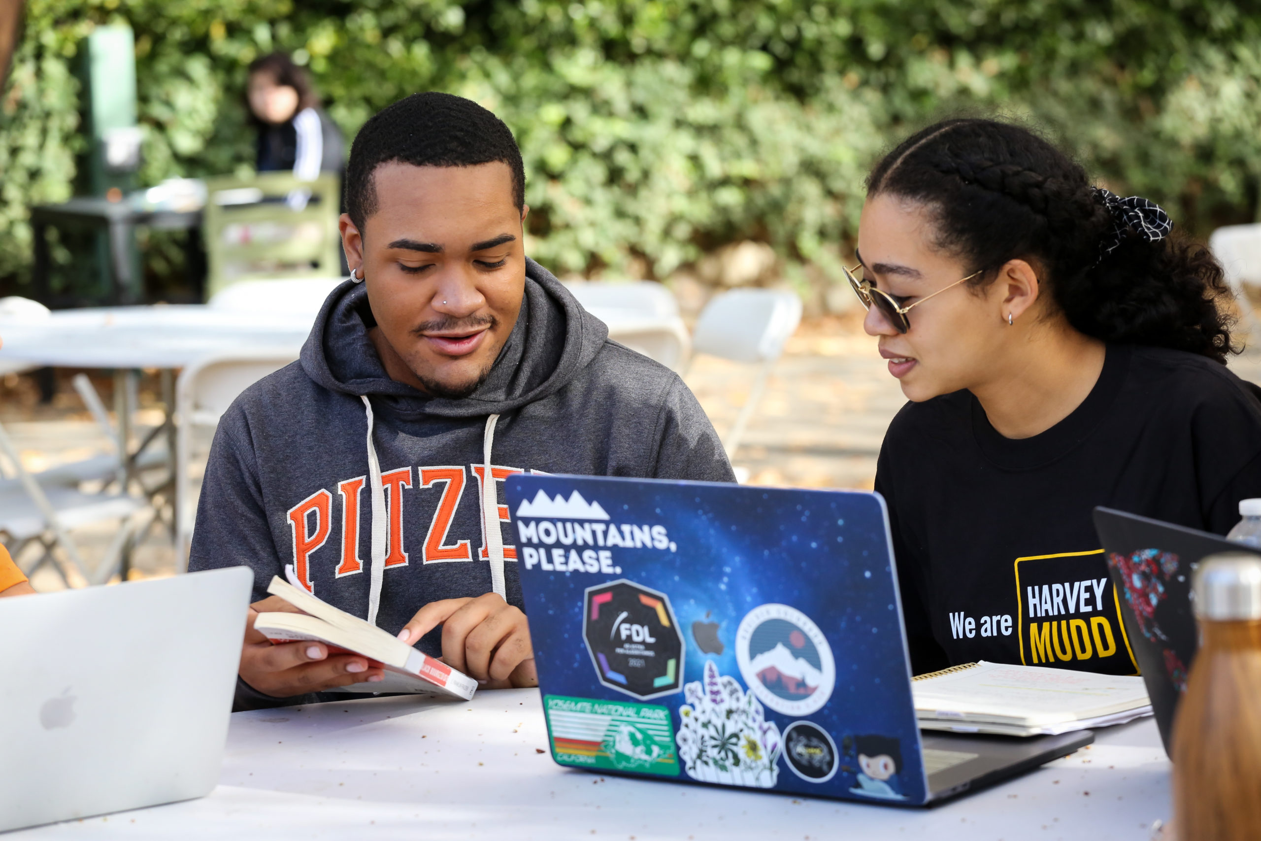 Two students sitting outdoors looking at book with laptops open.