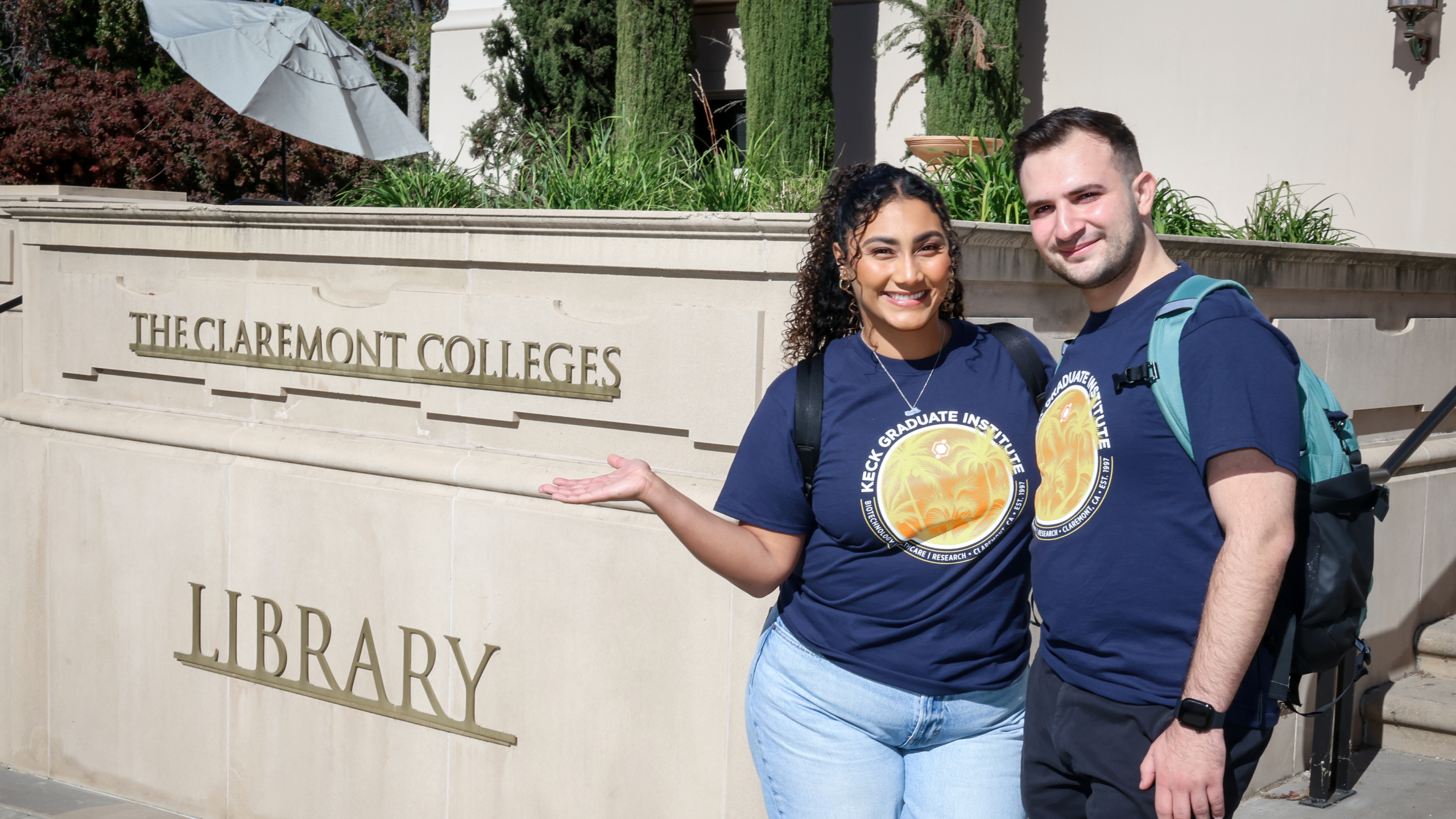 A female and male student standing and smiling in front of The Claremont Colleges Library sign
